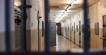 A view through prison bars looking down a long, empty corridor with cell doors.