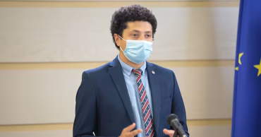 Tudor Bucșan wearing a suit and a face mask, speaking at a podium.