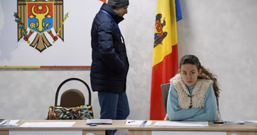 A woman sitting at a polling station table with Moldovan flags in the background.