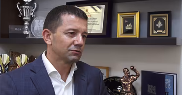 Dorin Damir in a suit, sitting in an office with sports trophies in the background.