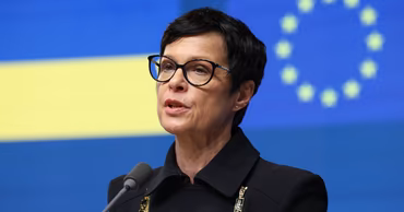 European Commissioner Marta Kos speaking at a press conference with EU flags in the background.