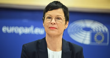 Marta Kos, European Commissioner, in a formal setting with the European Parliament logo behind her.