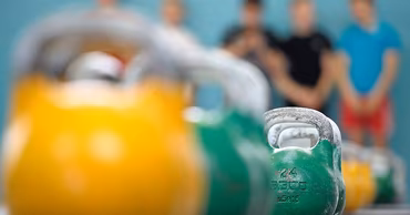 A row of colorful kettlebells in a gym setting with blurred athletes in the background.