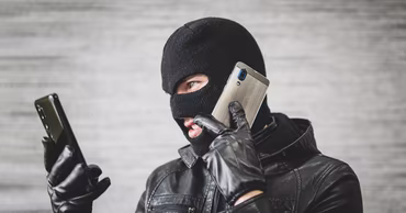 A person wearing a black balaclava and leather jacket holding two smartphones.