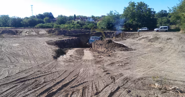 Construction machinery digging a large trench at a site with dirt and trees in the background.