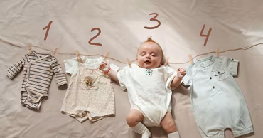 A baby lying on a bed next to baby clothes hanging on a string.