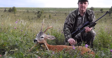 A hunter in camouflage gear posing with a hunted roe deer in a field.
