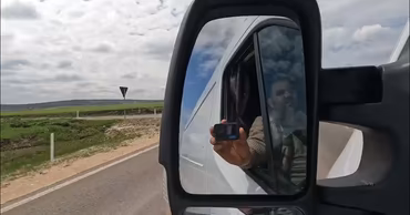 A view from a truck side mirror showing the driver holding a camera while driving on a road.