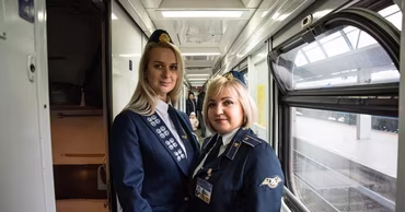 Two female train conductors in uniform standing inside a passenger train carriage.