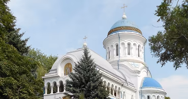 A white Orthodox church with blue domes surrounded by trees.