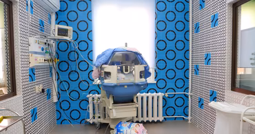 A neonatal incubator in a hospital room with blue patterned walls.