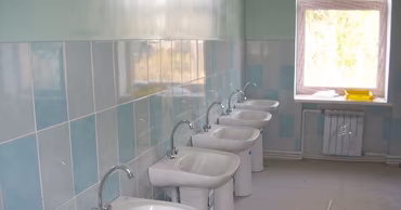 A row of new white ceramic washbasins installed in a school bathroom with tiled walls.