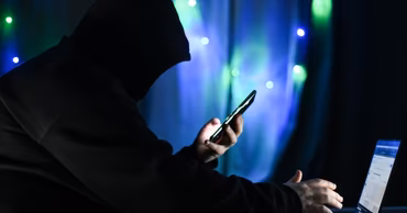 A person in a hoodie using a smartphone and laptop in a dark room.