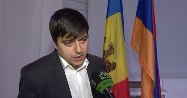 Gaik Vartanyan speaking into a microphone with Moldovan and Armenian flags in the background.