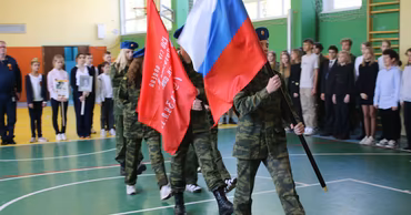Students in military-style uniforms carrying Russian and red flags in a school gymnasium.