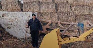 A farmer standing in front of hay bales and agricultural machinery.