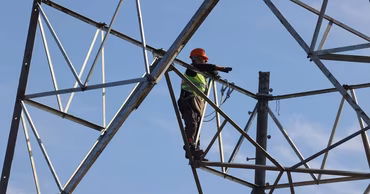 An electrician in safety gear working on a high-voltage power line tower against a blue sky.