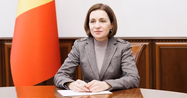Maia Sandu sitting at a table with the Moldovan flag in the background.