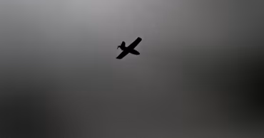 A silhouette of a drone flying against a grey, overcast sky.