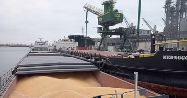 A barge loaded with grain docked at a port facility with loading cranes.
