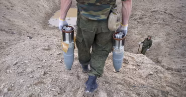 A soldier in camouflage carrying two large mortar shells in a sandy area.