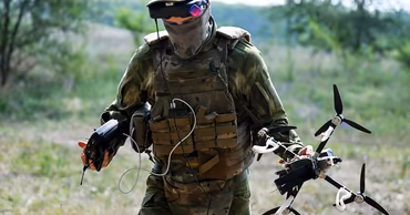A soldier in tactical gear holding a drone and a remote control in a field.