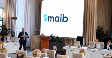 A speaker presenting at a maib corporate event with the bank's logo on a large screen.