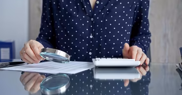 A person using a magnifying glass to examine financial documents on a desk.