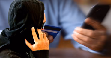 A person in a hoodie talking on a phone while holding a credit card.