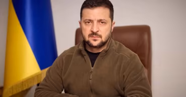 Volodymyr Zelenskyy sitting at a desk with a Ukrainian flag in the background.