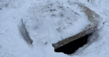 A snow-covered concrete well cover with an open gap in a winter setting.