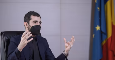 Claudiu Năsui wearing a suit and a face mask, gesturing while sitting at a table.