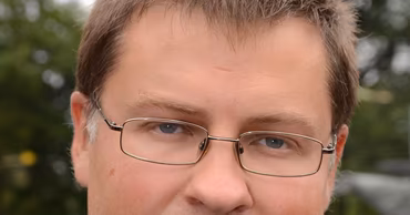 Portrait of Valdis Dombrovskis wearing a suit and glasses.