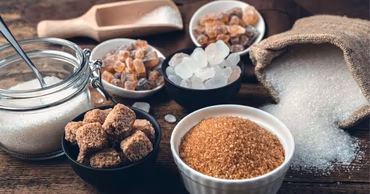 Various types of sugar in bowls and a burlap sack on a wooden table.