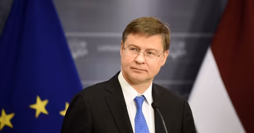 Valdis Dombrovskis speaking at a press conference with EU and Latvian flags in the background.
