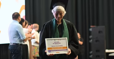 An elderly woman in graduation attire holding a diploma from the University of the Third Age.