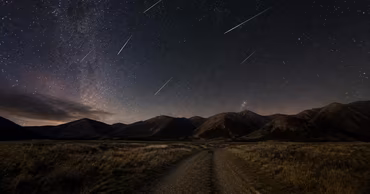 A night sky with visible meteor streaks over a dark landscape with mountains.