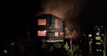 A wooden cottage burning at night with firefighters present at the scene.