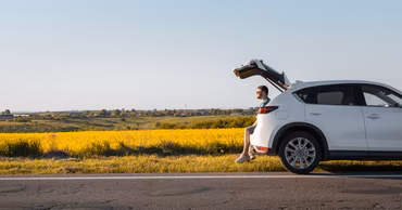 A man sitting in the open trunk of a white car parked by a field.