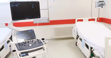 A modern ultrasound machine in a hospital room with a patient bed.