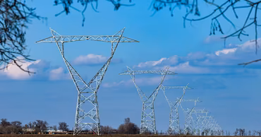 A row of high-voltage electricity pylons stretching across a field under a blue sky.
