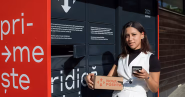 A woman holding a Nova Poshta parcel in front of a Nova Poshta automated locker.