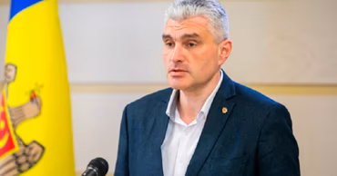 Alexandru Slusari speaking at a press conference with the Moldovan flag in the background.